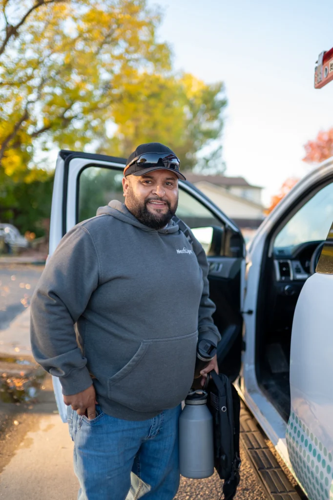 Smiling man by white truck in autumn suburb, golden hour, holding gear bag and canister.