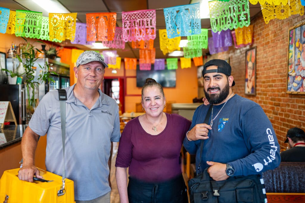 Smiling trio at colorful Mexican restaurant under papel picado banners; one holds yellow equipment case.