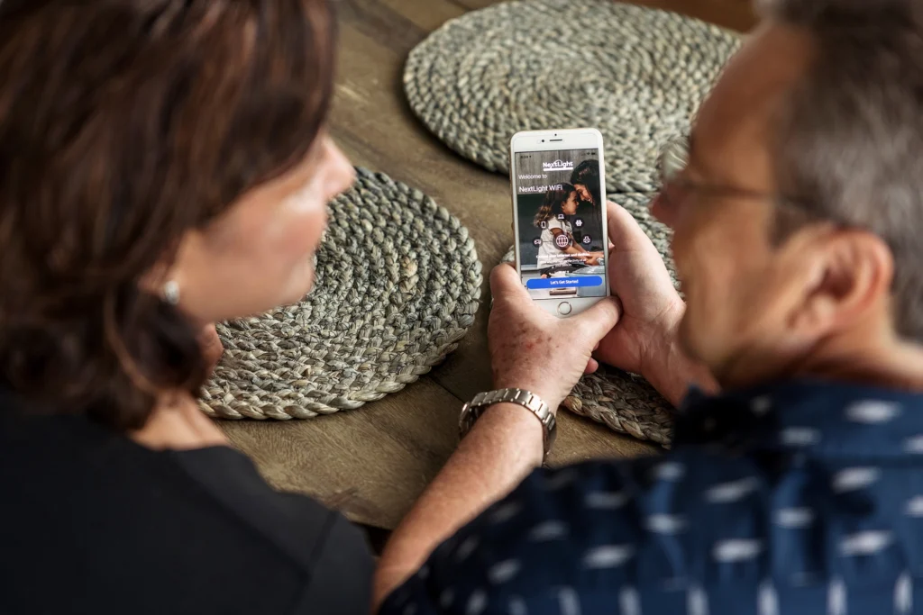 Two people at a table viewing a smartphone social media login screen with options to sign up and add a NextLight mesh WiFi satellite.